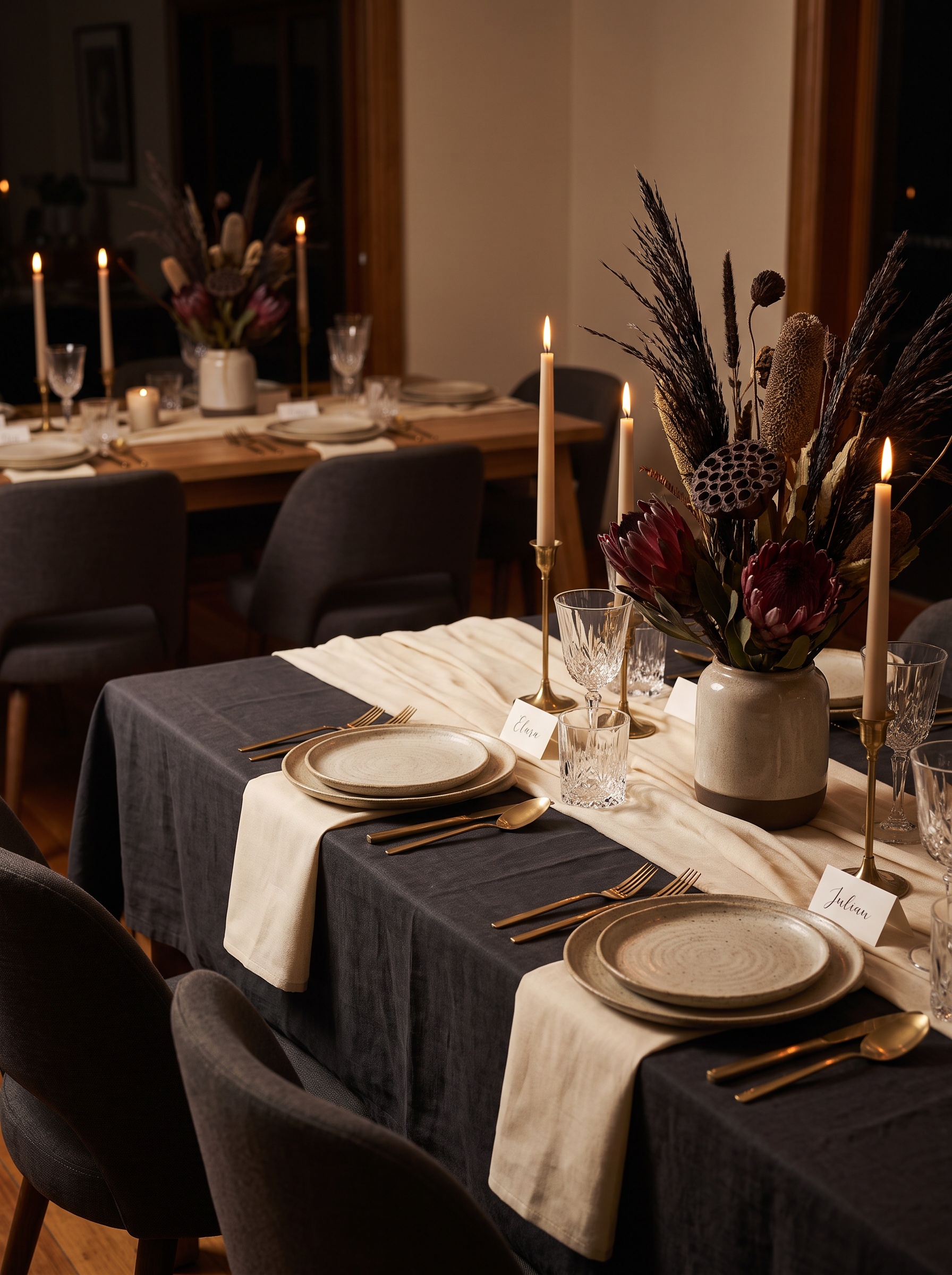 A candlelit table set with layered charcoal linen, brass candle holders, crystal glassware, and a sculptural botanical centerpiece, chairs empty